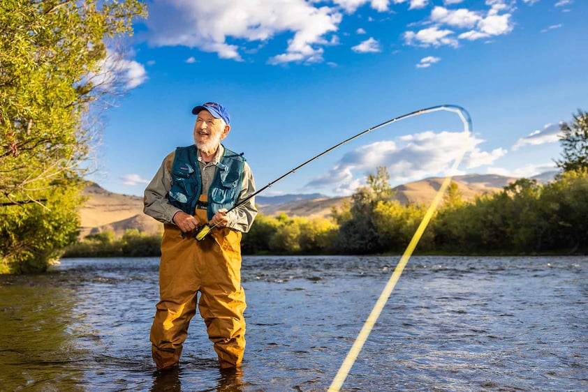Senior man in waders, a fishing vest and baseball cap standing in a river fishing