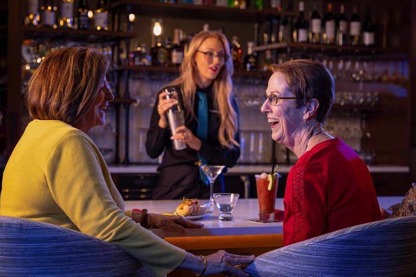 Two senior women with cocktails sitting at the bar with a female mixologist shaking a cocktail 