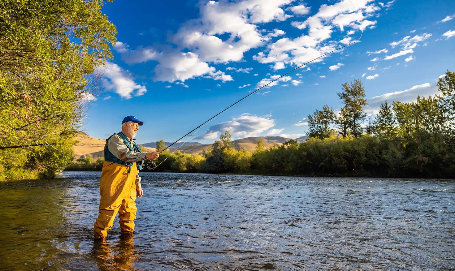 Senior man in waders, a fishing vest and baseball cap standing in a river fishing