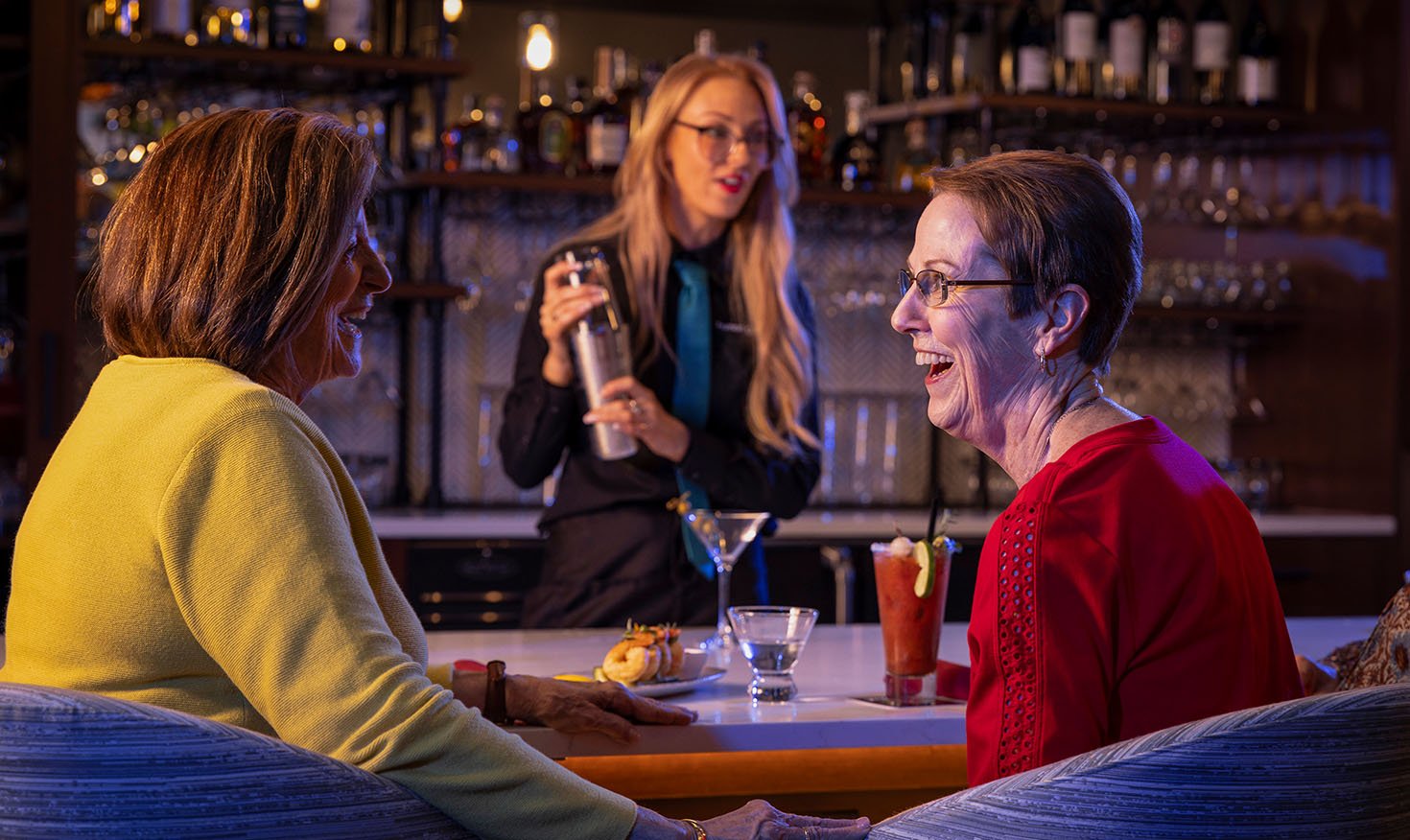 Two senior women with cocktails sitting at the bar with a female mixologist shaking a cocktail 