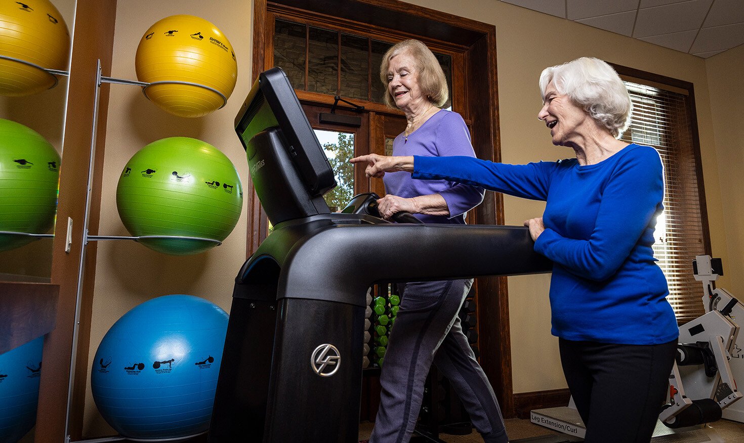 Senior woman standing and pointing at the display of a treadmill that another senior woman is walking on