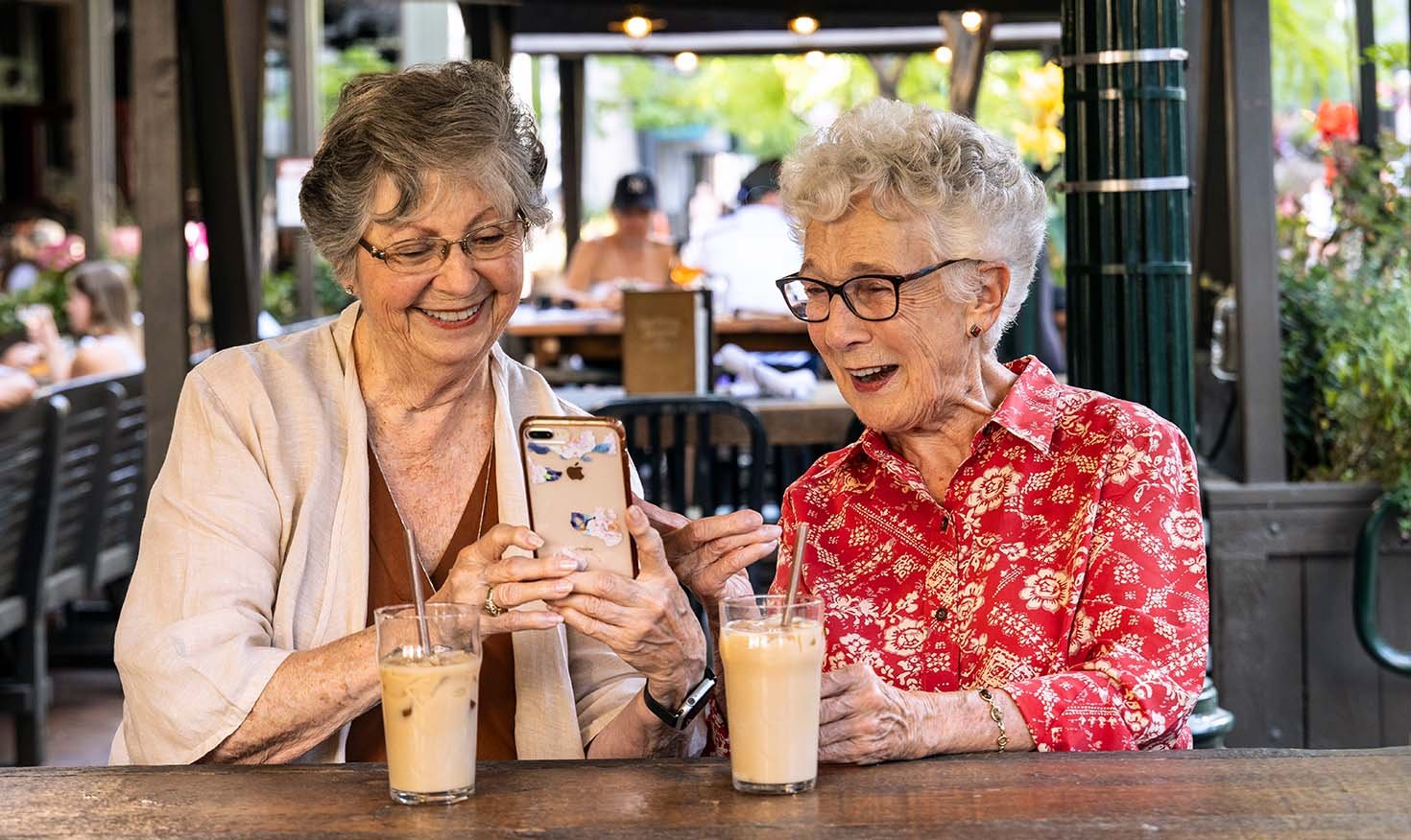 Two senior women sitting on a patio with iced coffee and looking at an iPhone