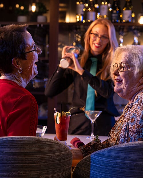 Two senior women with cocktails sitting at the bar with a female mixologist shaking a cocktail 
