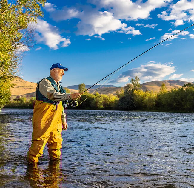 Senior man in waders, a fishing vest and baseball cap standing in a river fishing