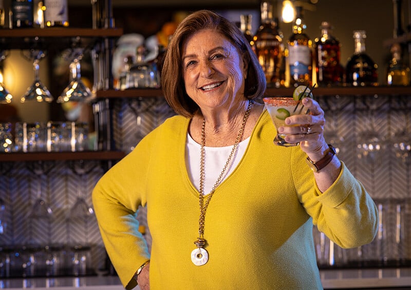 Senior woman in front of a bar holding up a cocktail