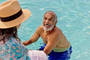 Senior man in the pool smiling at a senior woman wearing a sun hat