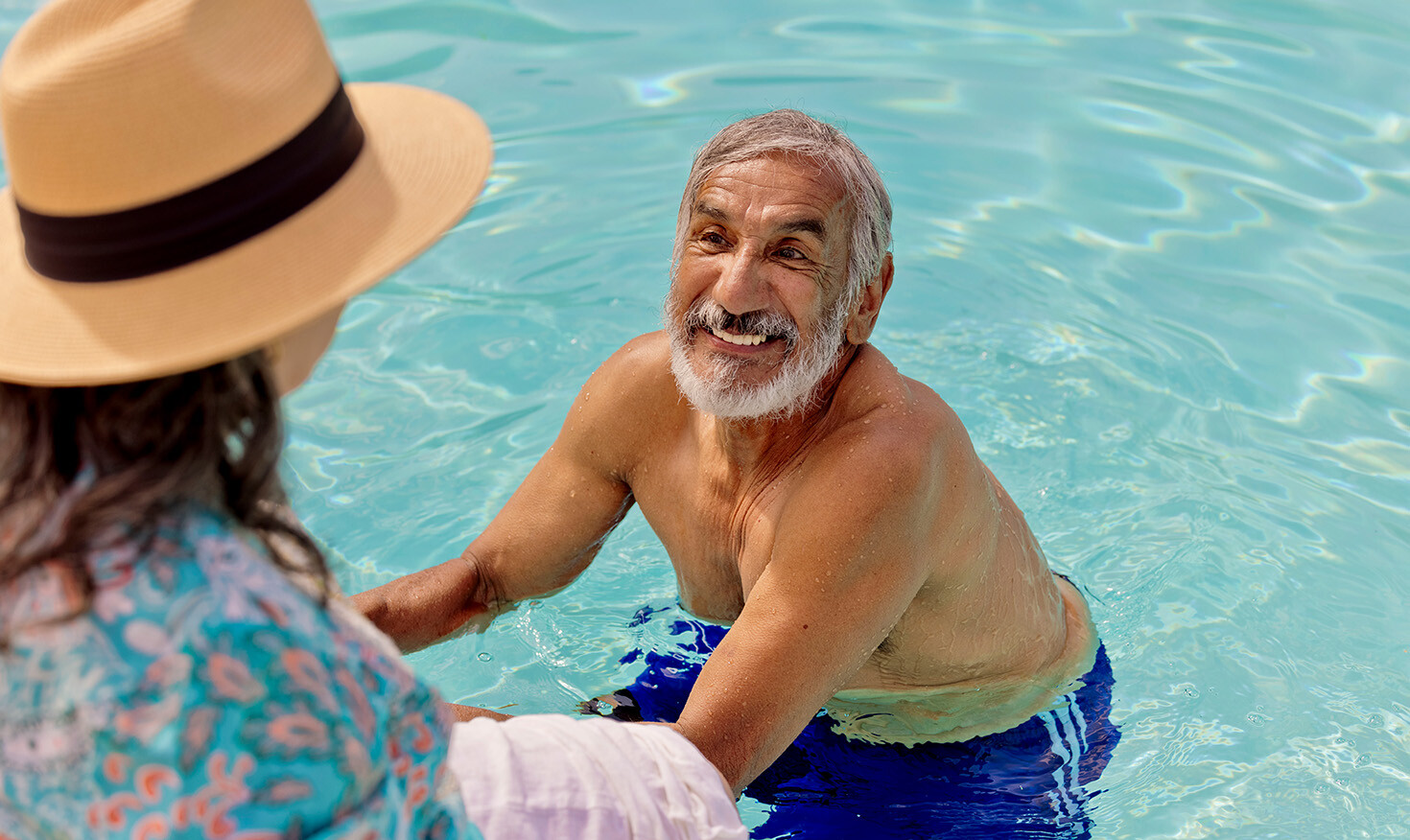 Senior man in the pool smiling at a senior woman wearing a sun hat