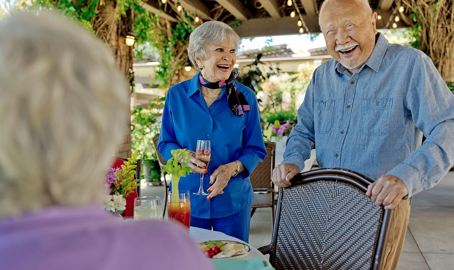 Smiling senior man and woman standing and talking to a senior woman sitting at an outdoor dining table