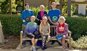 Group of six seniors, three sitting on a bench and three standing behind it, holding pickleball paddles