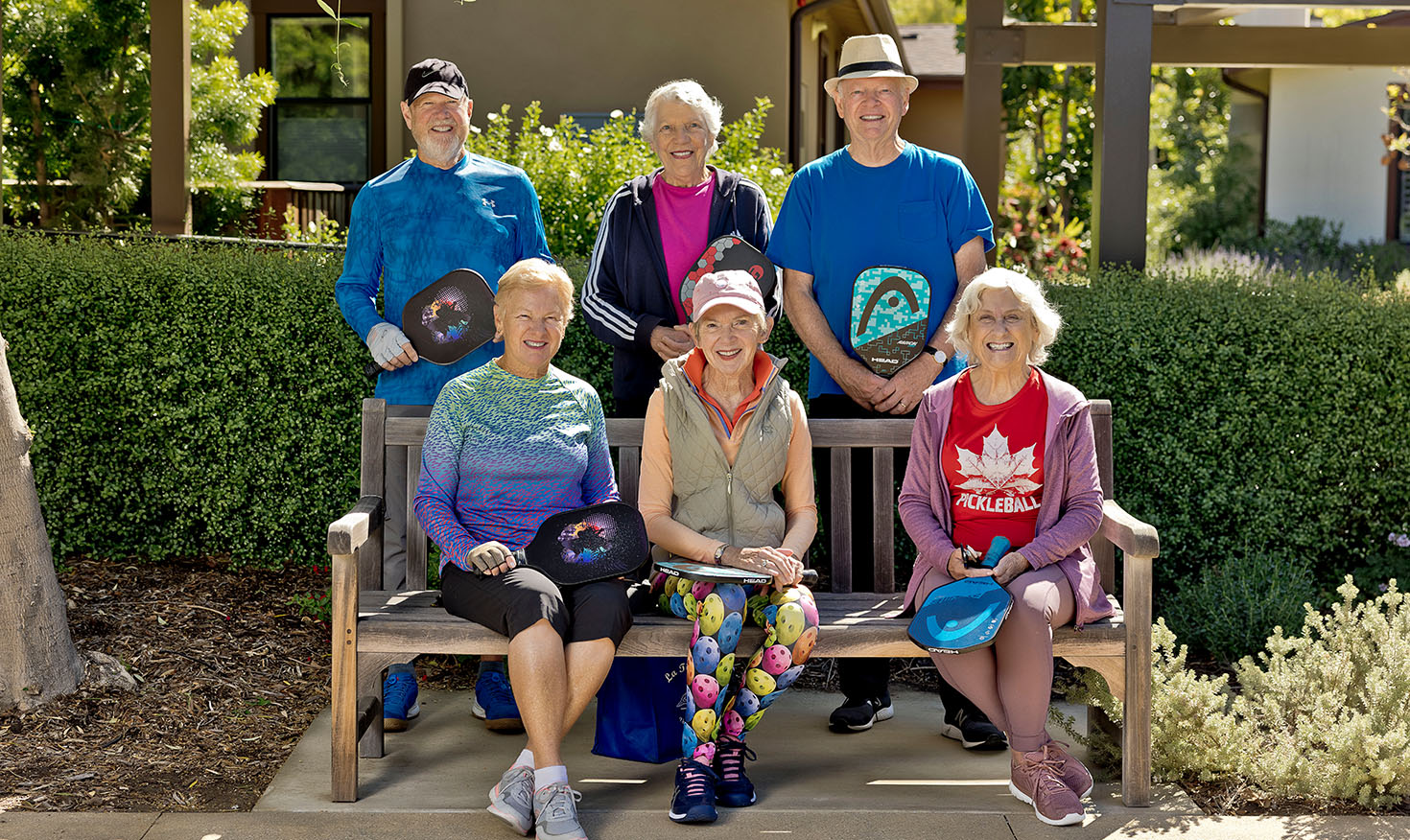 Group of six seniors, three sitting on a bench and three standing behind it, holding pickleball paddles