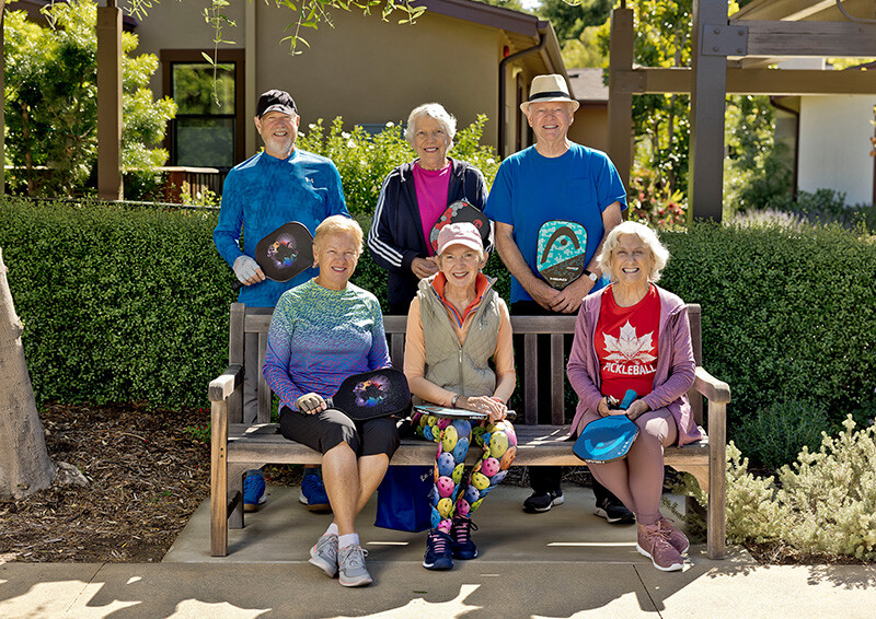 Group of six seniors, three sitting on a bench and three standing behind it, holding pickleball paddles