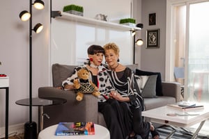 Two women sit closely on a grey sofa in a stylish living room, holding a small stuffed toy. They wear patterned black-and-white outfits.
