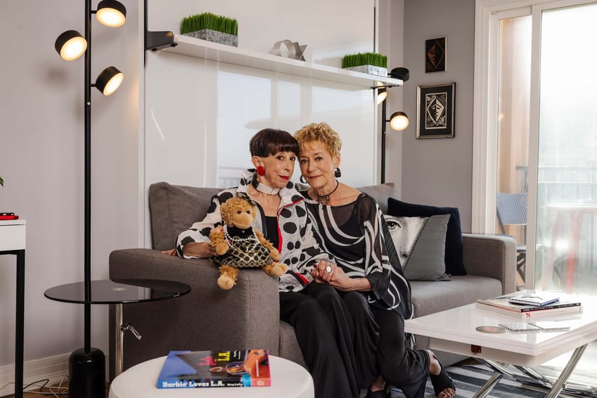 Two women sit closely on a grey sofa in a stylish living room, holding a small stuffed toy. They wear patterned black-and-white outfits.