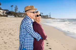 Senior couple smiling on a sunny beach with ocean waves and houses in the background.