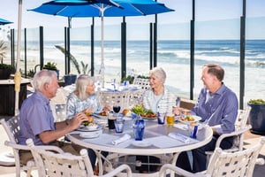 Four older adults dining at an outdoor beachside restaurant with a view of the ocean.