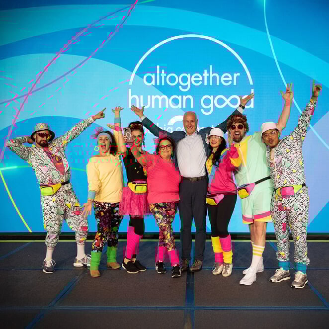 A group of eight people in colorful, retro outfits pose joyfully on stage. Behind them, a vibrant blue background says, altogether human good.