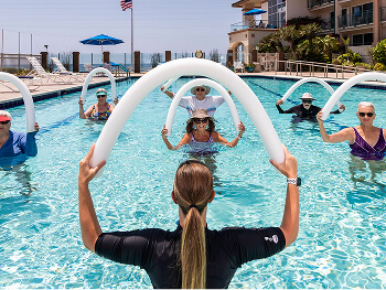A group of six adults engages in an aquatic exercise class, holding pool noodles in a rooftop pool