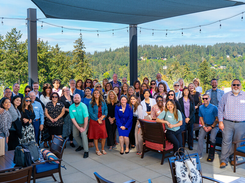 A large group of people pose smiling on a sunny deck with a forested backdrop.