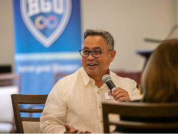 A man in a white barong speaks into a microphone, smiling warmly at an indoor event. A blue and white banner is slightly blurred in the background.