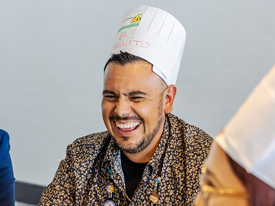 A man wearing a patterned shirt and a paper chef's hat laughs heartily, creating a joyful and lighthearted atmosphere in the room.