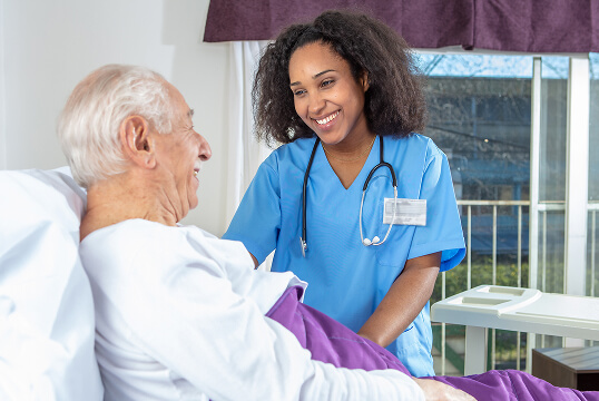 A nurse in blue scrubs, smiling warmly, attends to an elderly patient in a hospital bed. The atmosphere is caring and supportive, with natural light streaming in.