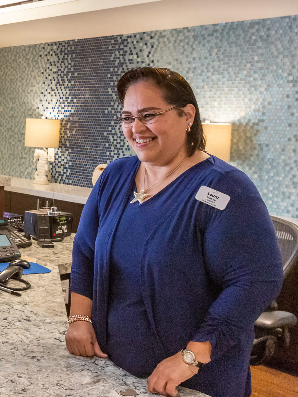 A receptionist in a blue top warmly greets two older women at a modern, well-lit front desk, creating a welcoming and friendly atmosphere.