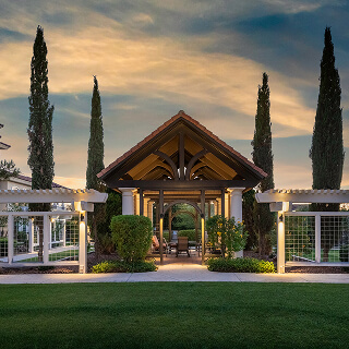 A serene garden pavilion at dusk, framed by tall cypress trees and pergolas, with a warm, inviting glow against a soft, cloudy sky.