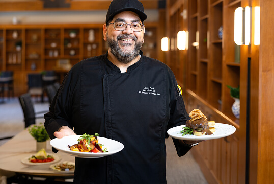 A smiling chef in a black uniform holds two plated dishes, featuring a vibrant salad and a gourmet entrée, in a warmly lit dining setting.