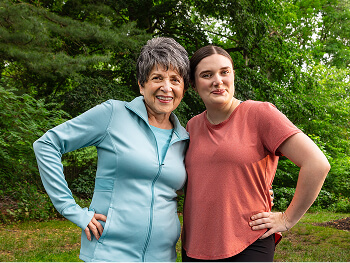 A smiling senior woman in a light blue jacket and a young woman in a red shirt stand outdoors, posing confidently with their hands on their hips, surrounded by lush greenery