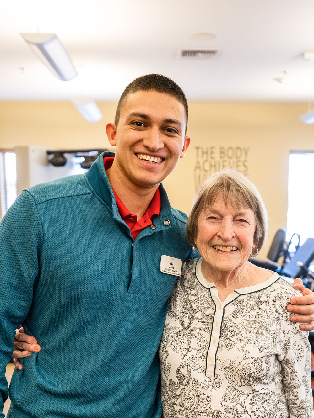 A smiling young man in a teal sweater and an elderly woman in a patterned blouse pose together in a warmly lit room, conveying a sense of joy and connection.
