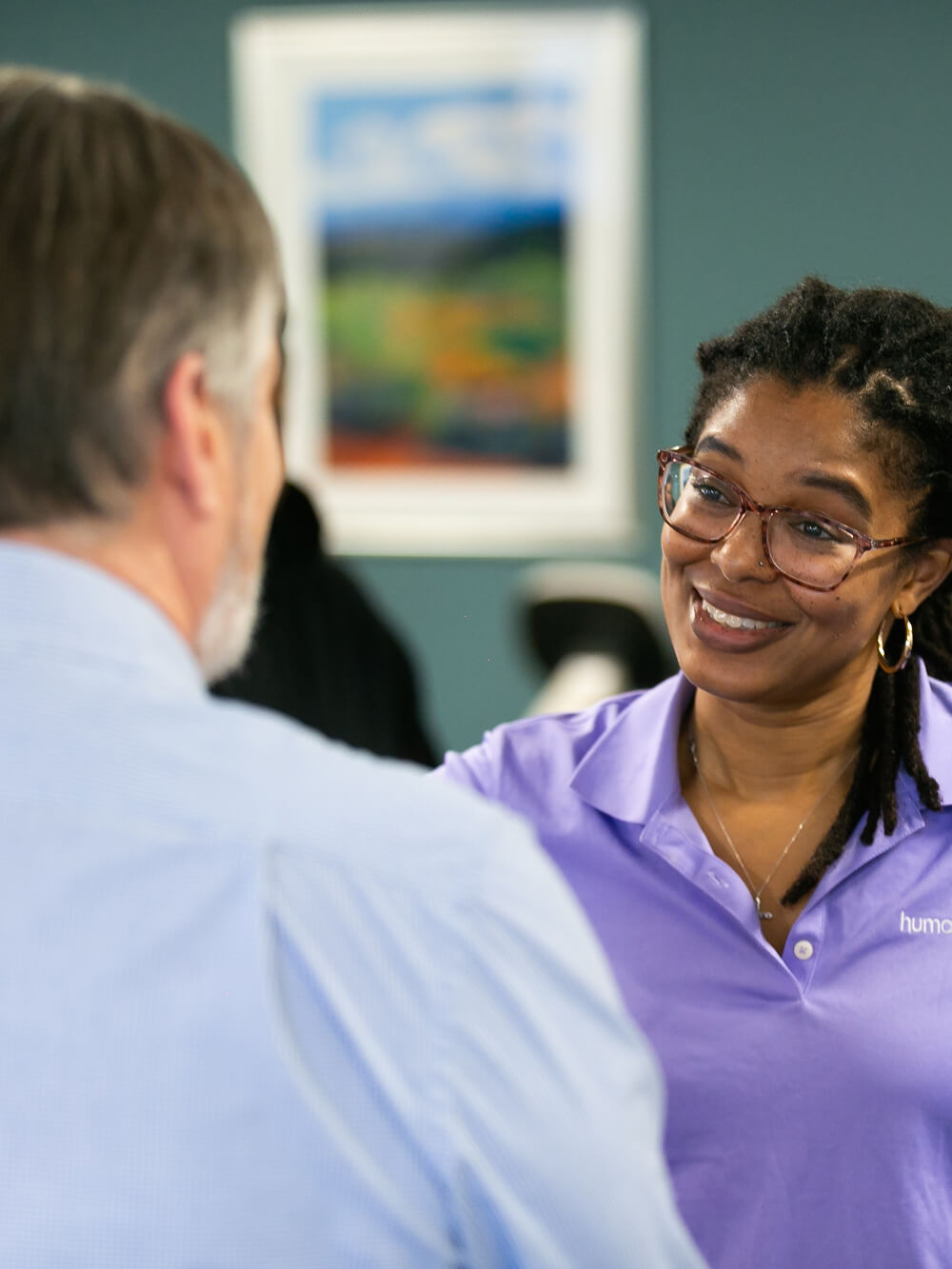 A woman in a purple shirt and glasses smiles as she talks to a man in a blue shirt at a social gathering.