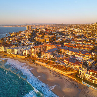 Aerial view of a coastal city at sunset, featuring sandy beaches, waves, and a blend of modern and historic buildings glowing in warm golden light.