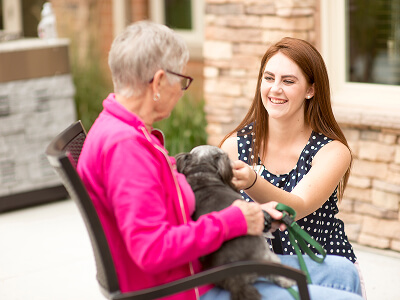 An elderly woman in a pink jacket holds a small dog while chatting outside with a young woman in a blue polka dot top, both smiling warmly.