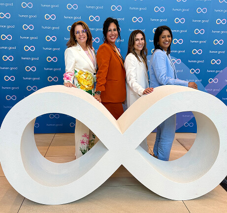 Four women in vibrant suits pose smiling behind a large white infinity symbol.