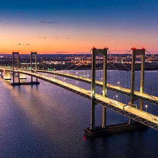 Illuminated suspension bridge at dusk spans calm blue waters, with city lights twinkling in the background under a purple-orange sunset sky.