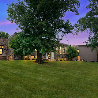 Large building with numerous windows, warmly lit inside, viewed from a grassy lawn at dusk. A tall tree stands prominently, with a purple and blue sky above.