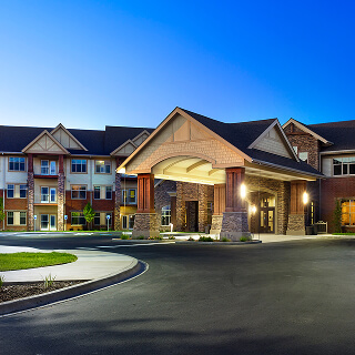 Modern apartment building at dusk with warm exterior lighting, featuring a covered entrance and manicured landscaping against a clear blue sky.