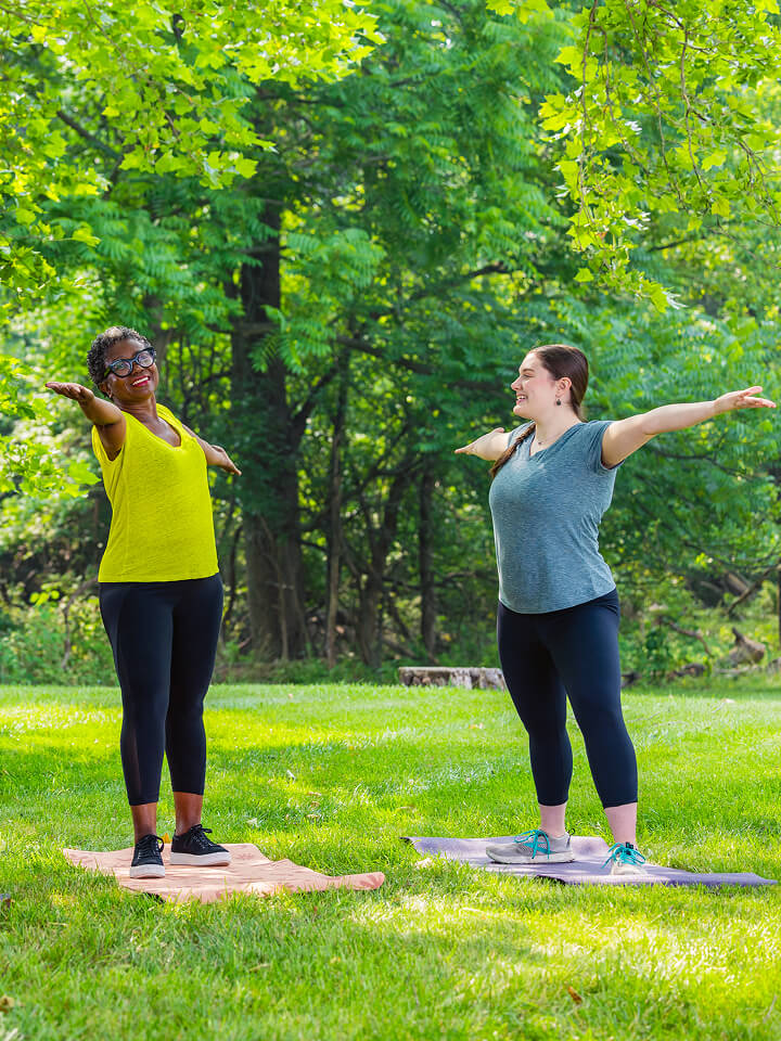 Two women practice yoga on mats in a lush park, smiling with outstretched arms.
