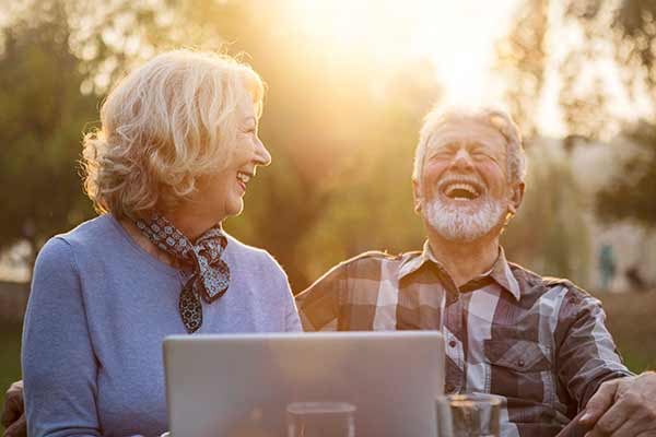 Senior couple sitting outdoors with a laptop, laughing together in the warm glow of the sunset