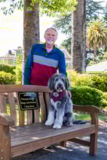Senior man standing behind an outdoor bench with a dog sitting on it at Plymouth Village