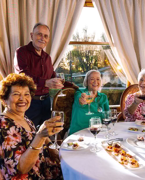 Four senior friends in the Plymouth Village dining room holding up glasses of wine