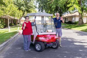 Senior man and woman standing next to a red golf cart and waving on the grounds of Plymouth Villag