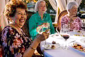 Three senior women sitting at a dining table holding up glasses of wine