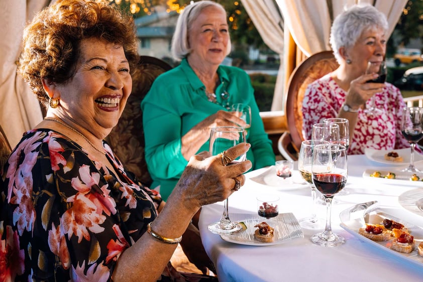Three senior women sitting at a dining table holding up glasses of wine