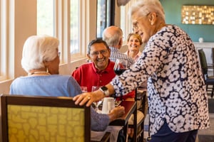 Senior woman stopping by a table in the Plymouth Village dining room to chat with a senior couple enjoying a meal