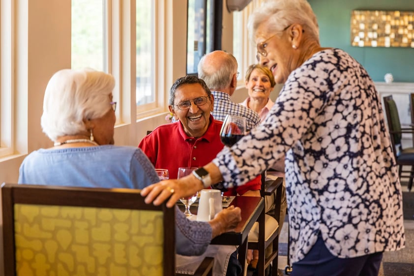 Senior woman stopping by a table in the Plymouth Village dining room to chat with a senior couple enjoying a meal