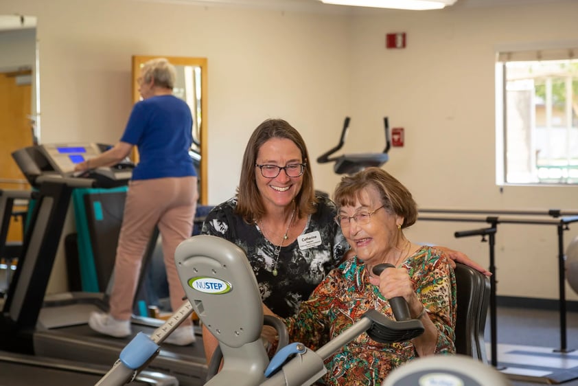 Female fitness trainer next to a senior woman using a recumbent bike in the Plymouth Village fitness center