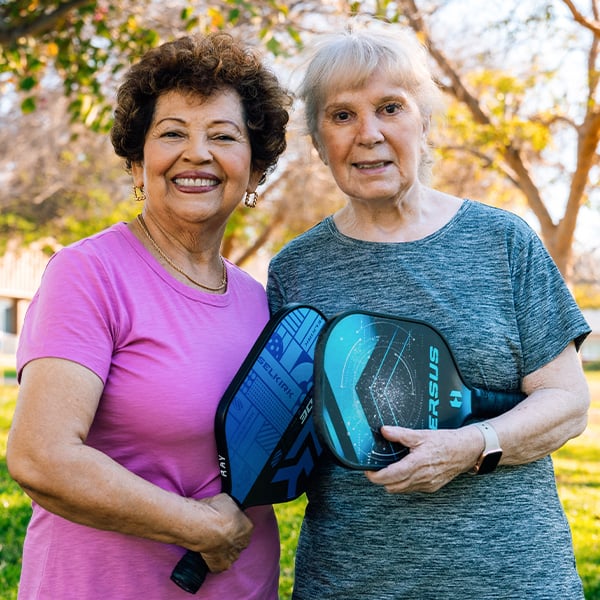 Two smiling senior women outdoors holding pickleball paddles
