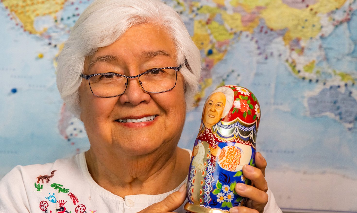 Janet, a senior resident of Plymouth Village, in front of a map of the world holding up a Russian nesting doll souvenir