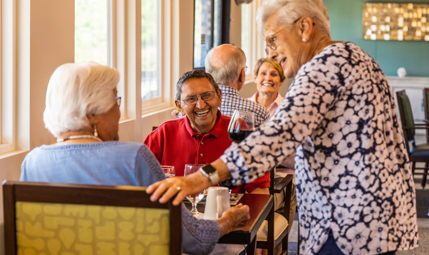 Senior woman stopping by a table in the Plymouth Village dining room to chat with a senior couple enjoying a meal
